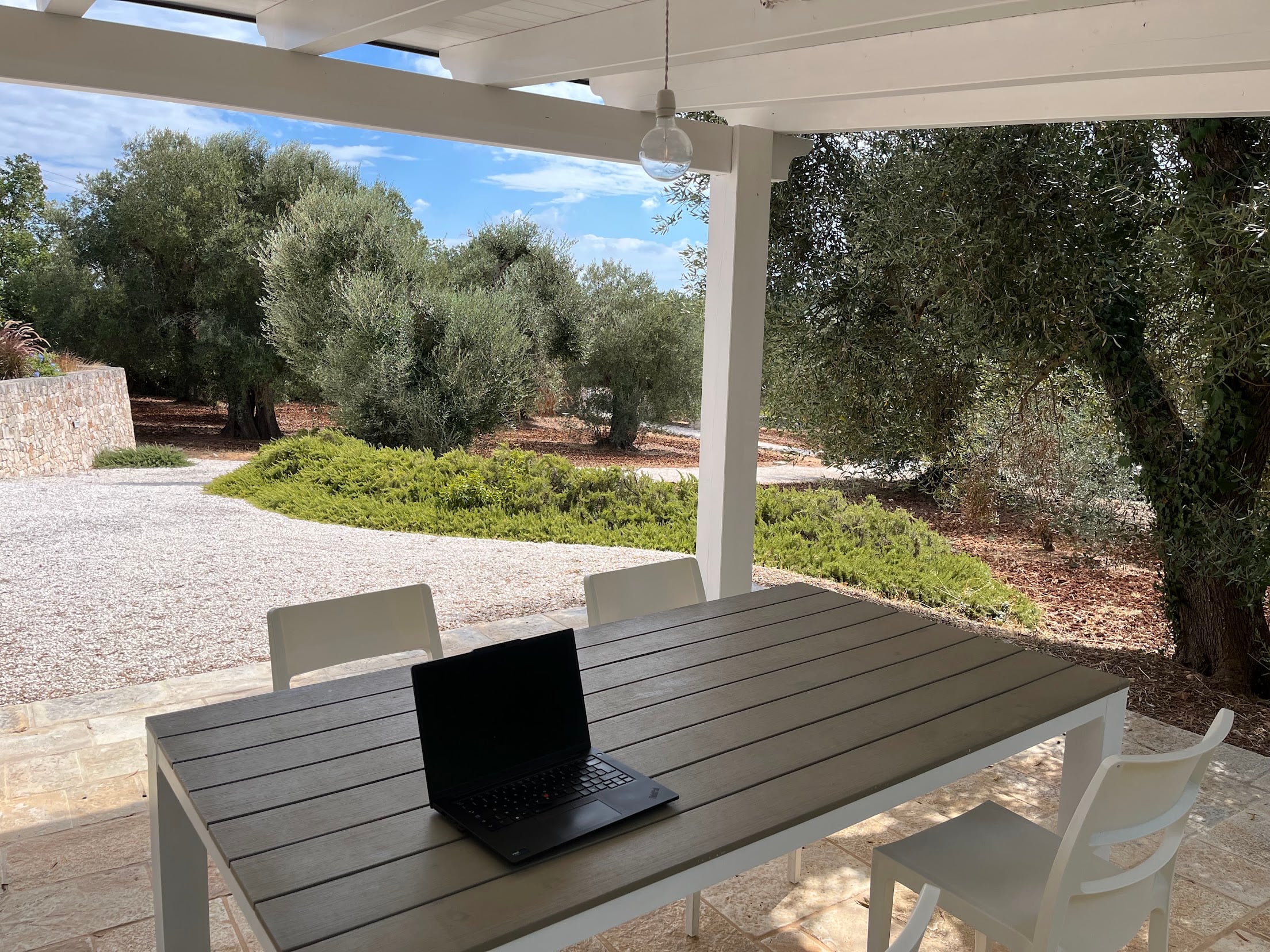 A laptop open on a garden table under a covered veranda, looking out over gravel and an olive grove